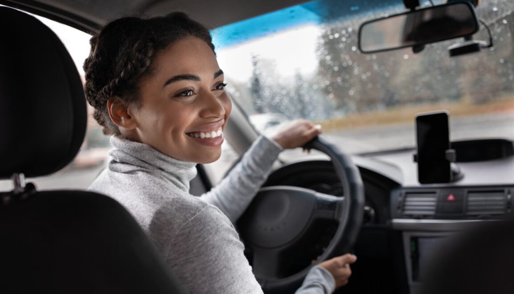 Excellent trip, auto travel and enjoy journey in town. Happy young african american woman driver turns to passenger in back seat. View of city road, window with raindrops and smartphone, copy space